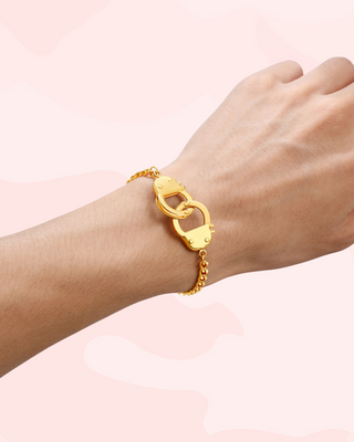 Gold bracelet with a unique handcuff clasp design on a wrist against a light pink background