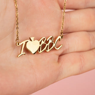 A gold "I Heart BBC" necklace with bold lettering and a heart symbol is elegantly draped across an open hand. The delicate chain cascades naturally over the fingers, catching the light and highlighting its polished shine