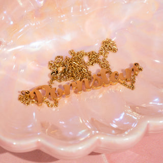 The gold "Pornstar" necklace with elegant, cursive lettering rests delicately on a glossy pink surface.
