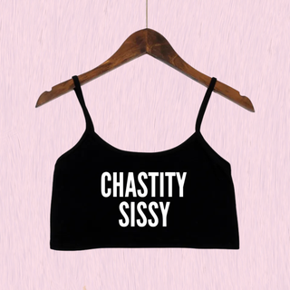 A black spaghetti-strap crop top with the words 'CHASTITY SISSY' printed in white block letters, hanging on a wooden hanger against a light pink textured background.