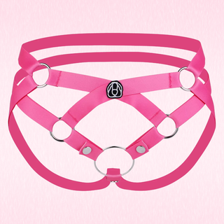 A hot pink strappy harness-style chastity belt with multiple circular metal rings and a small black-and-white emblem at the center, shown on a pale pink background.