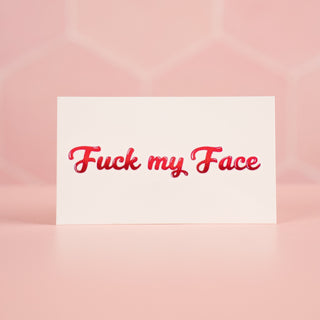 A close up of the Fuck my Face temporary tattoo, the lettering is bright pink and is shown on its white backing paper.