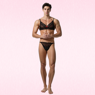 A male model wearing  Luna Lace Duo black lace lingerie set, consisting of a bra and matching briefs, standing against a light pink textured background.