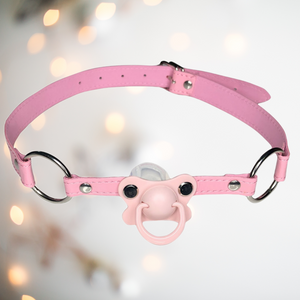 A pink pvc long gag that has a pale pink dummy fixed to the front which the wearer will have to hold in their mouth and a  silver buckle fastening to the back.